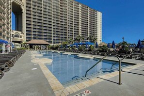 Ein Hotelpoolbereich mit einem großen Gebäude im Hintergrund, Liegestühlen und Sonnenschirmen.