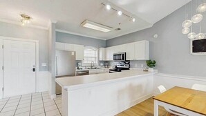 A modern kitchen with white cabinetry, a central island, and a dining area with a wooden table. - Carnation Unit 302 Condo (Myrtle Beach)