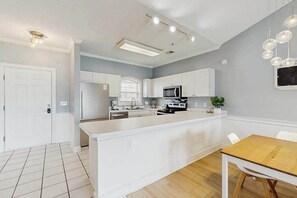 A modern kitchen with white cabinetry, a central island, and a dining area with a wooden table. - Carnation Unit 302 Condo (Myrtle Beach)