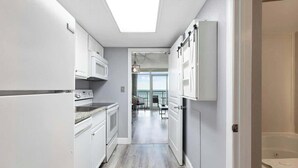 A narrow kitchen with white cabinets, a built-in oven, and a microwave. There is a skylight above the sink, and the kitchen leads to a dining area with a view of the outdoors. - Bay Watch Resort 0439 (North Myrtle Beach)