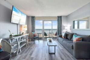 A modern living room with a sofa, a coffee table, a TV mounted on the wall, and a balcony with a view. - Bay Watch Resort 0439 (North Myrtle Beach)