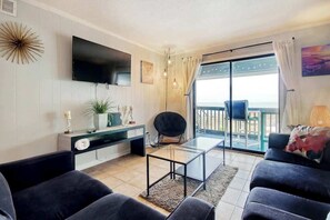 A modern living room with a flat-screen TV, a glass coffee table, and a sofa. There is a balcony with a view. - Cherry Grove Retreat (North Myrtle Beach)