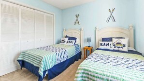 Un dormitorio con dos camas, cada una con una colcha estampada y almohadas con texto. Hay dos elementos montados en la pared que parecen murciélagos cruzados, y un armario blanco con persianas horizontales.