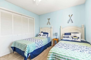 A bedroom with two beds, each with a patterned bedspread and pillows with text. There are two wall-mounted items resembling crossed bats, and a white closet with horizontal blinds. - Coastal Comforts Retreat (Surfside Beach)
