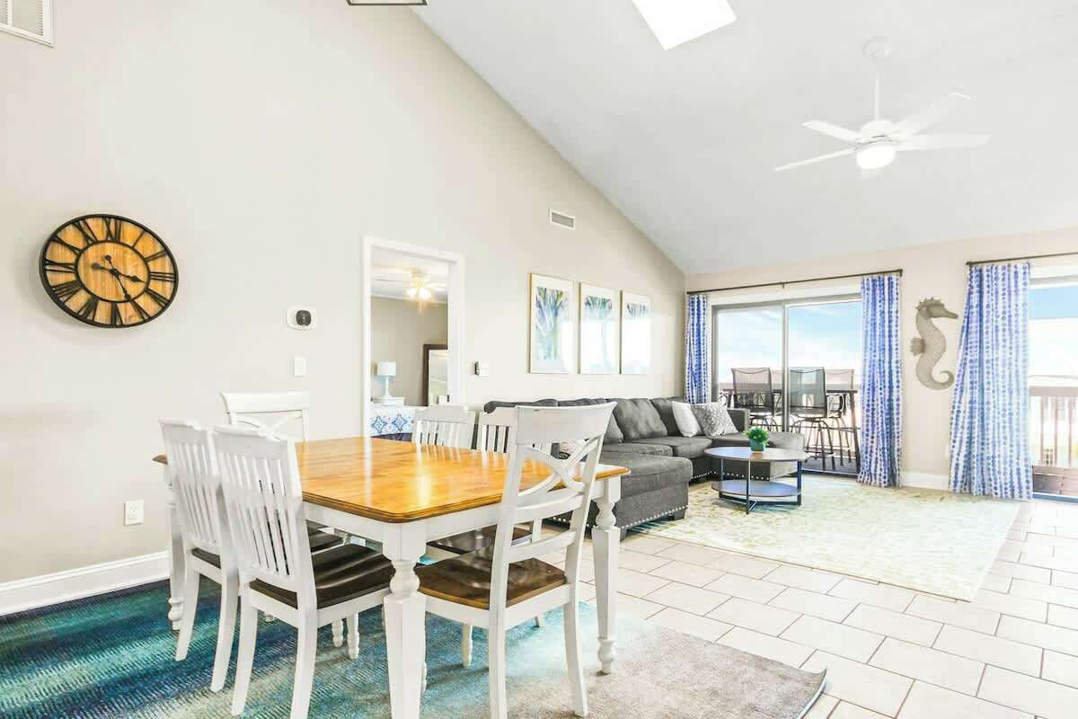 A spacious living area with a dining table, white chairs, a sofa, and a large window with curtains.