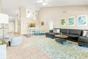 A spacious living area with a gray sofa, a coffee table, and a patterned rug. There is a dining area with white chairs and a table in the background. - Coastal Comforts Retreat (Surfside Beach)