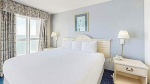 A hotel room with a large bed, two bedside lamps, a nightstand, and a window with curtains. - Bay Watch Resort 1637 (North Myrtle Beach)