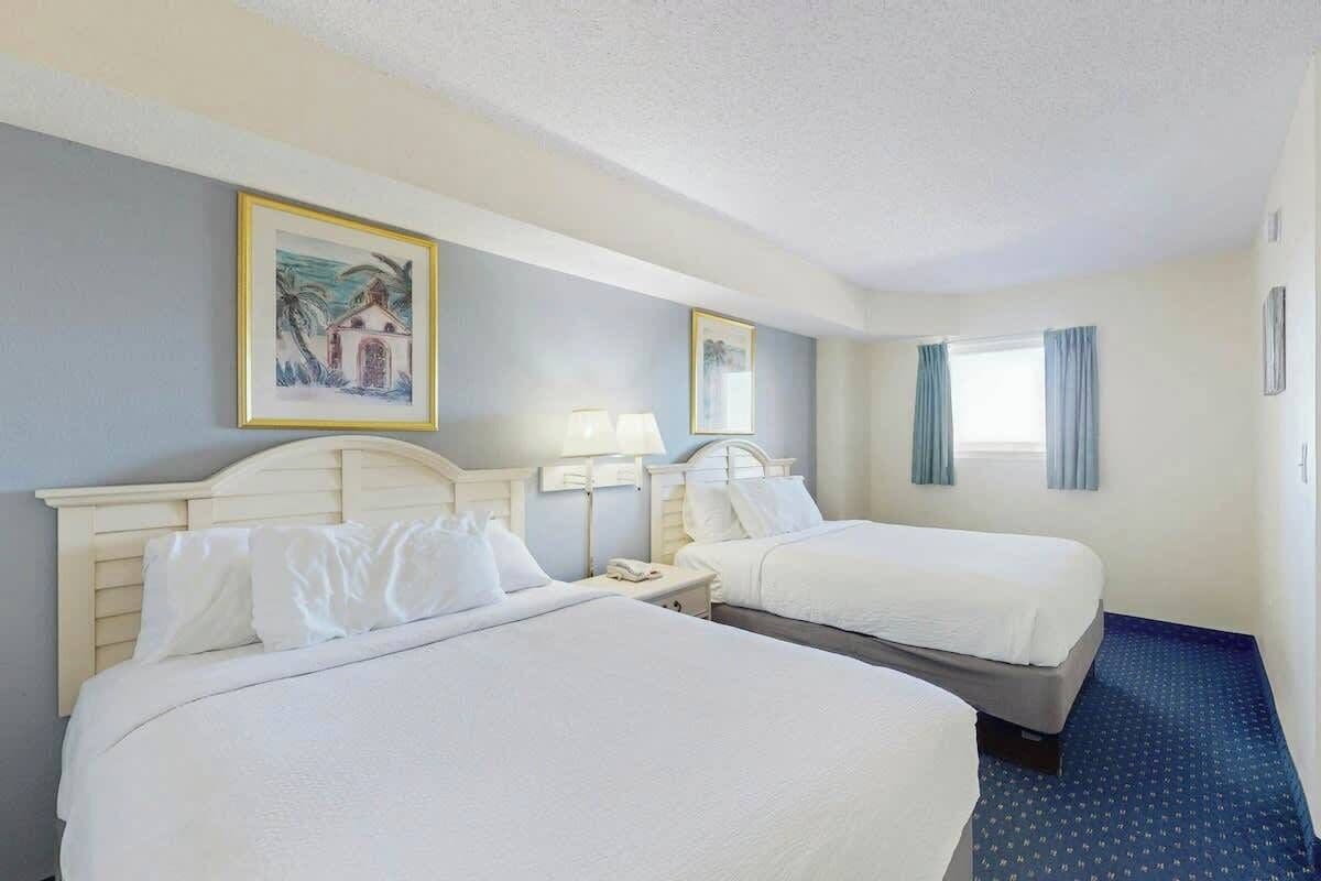 Quarto de hotel com duas camas, uma mesa de cabeceira, uma luminária e uma janela com cortinas.