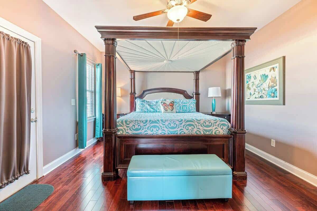 A bedroom with a four-poster bed, a ceiling fan, a window with curtains, and a framed picture on the wall.