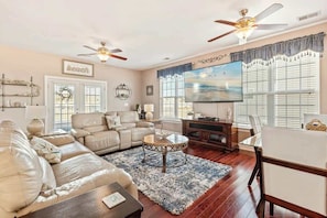 A living room with a beige sofa, a coffee table, and a TV. There are ceiling fans, a fireplace, and a window with blinds. - Waterfront Galore Beach Home (North Myrtle Beach)