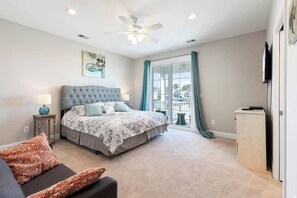 A bedroom with a large bed, a ceiling fan, a window with curtains, and a TV on the wall. - Tidewatch Charms Townhouse (North Myrtle Beach)