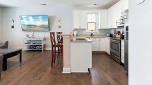 A modern kitchen with white cabinetry, a central island, and a dining area with chairs. - Tidewatch Charms Townhouse (North Myrtle Beach)