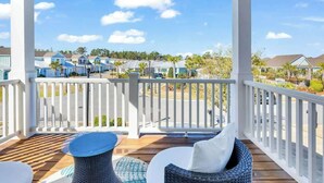A covered outdoor area with a wooden deck, white railings, wicker furniture, and a view of a residential area with houses and greenery. - Tidewatch Charms Townhouse (North Myrtle Beach)