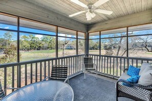 A covered outdoor seating area with a table and chairs, a sofa with cushions, and a view of a grassy field and trees. - Beaches and Birdies Retreat (North Myrtle Beach)