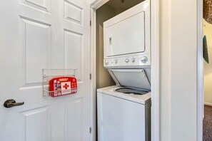 A compact laundry room with a washing machine and dryer, a white door, and a red emergency kit on a wall-mounted rack. - Beaches and Birdies Retreat (North Myrtle Beach)