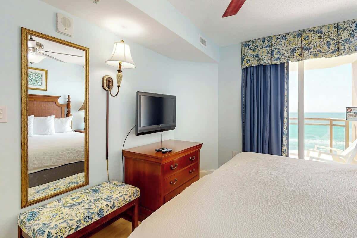 Une chambre d’hôtel équipée d’un lit, d’une télévision, d’un miroir, d’une table de chevet et d’une fenêtre avec des rideaux.