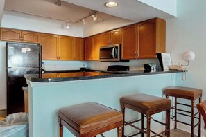 A kitchen with wooden cabinets, a refrigerator, a microwave, and a bar area with stools. - Bahama Sands Unit 0508 (North Myrtle Beach)