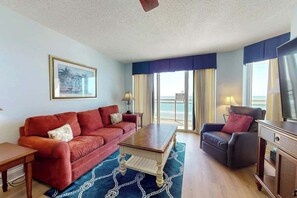 A living room with a red sofa, a wooden coffee table, and a grey armchair. There is a television on a wooden stand, and a framed picture on the wall. - Bahama Sands Unit 0508 (North Myrtle Beach)
