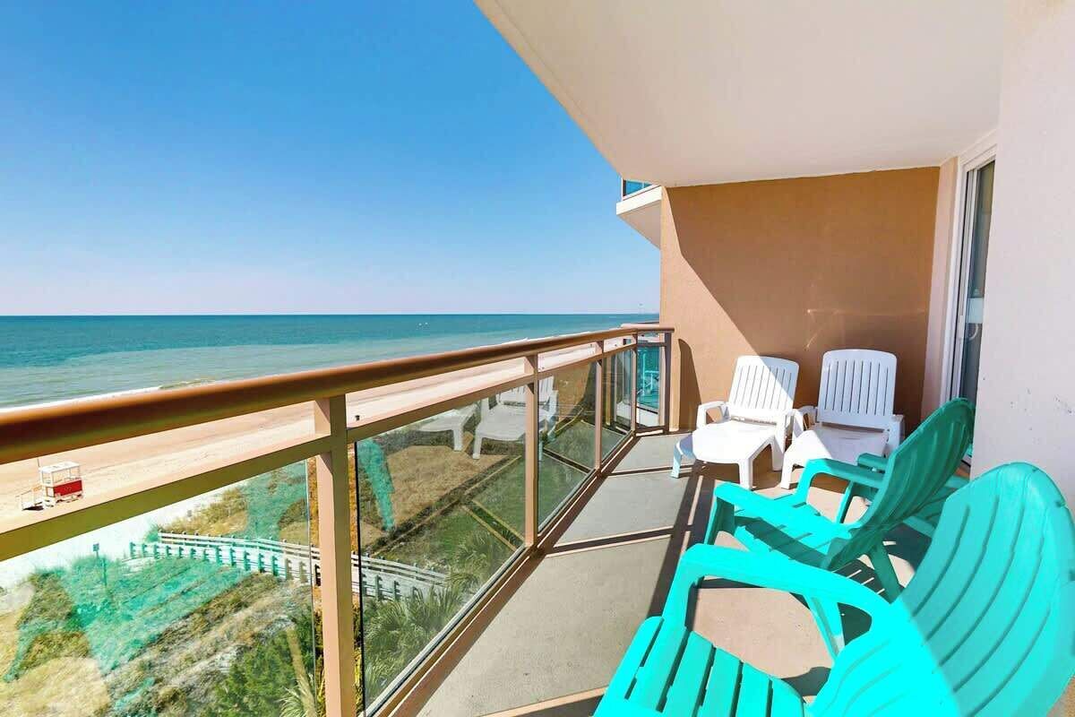 Un balcon avec vue sur la plage et l’océan, aménagé avec des chaises longues turquoise et des chaises blanches.