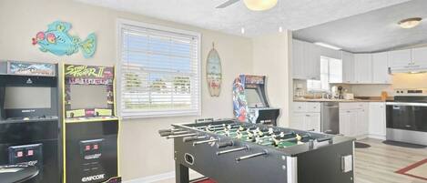 A room with a foosball table, arcade games, a ceiling fan, and a kitchen area.