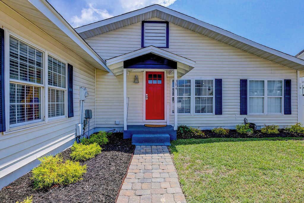 Une maison de plain-pied avec une porte d’entrée rouge, un petit porche et un chemin en briques menant à l’entrée.