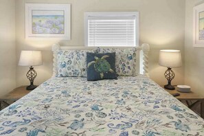 Un dormitorio con una colcha de estampado floral, dos lámparas de noche, un ventanal con persianas y cuadros en las paredes.