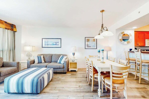 A modern living room with a sofa, ottoman, and dining table with chairs. There is a kitchen area with cabinets and a refrigerator. - Harbourgate 0526 - Serenity on The Marina (North Myrtle Beach)