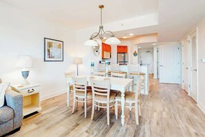 A modern kitchen with white cabinets, a stainless steel refrigerator, and a dining area with a white table and chairs. - Harbourgate 0526 - Serenity on The Marina (North Myrtle Beach)