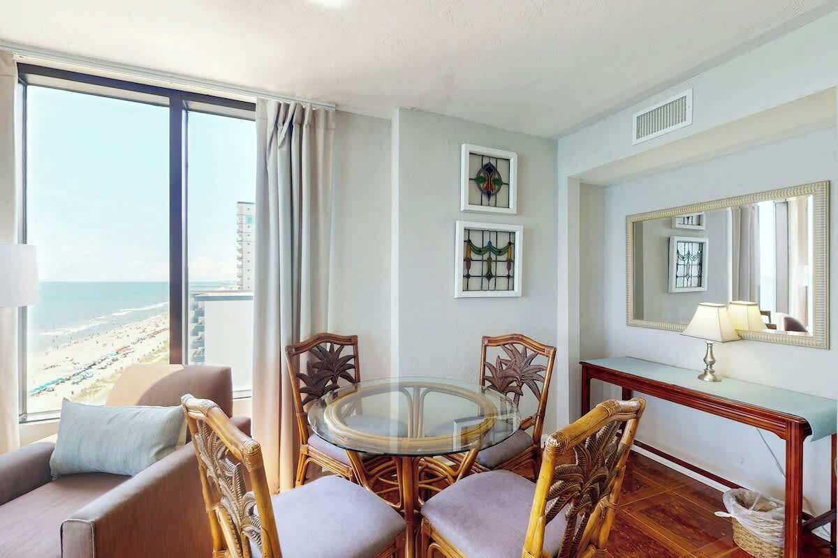 Um quarto com uma mesa de vidro redonda, cadeiras de vime, um sofá, um aparador com uma luminária e um espelho grande.