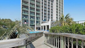 Eine Holzterrasse führt zu einem Poolbereich, im Hintergrund ist ein Hochhaus-Hotel zu sehen.