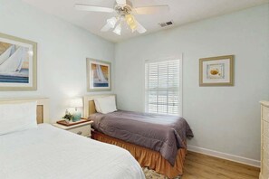 Ein Schlafzimmer mit zwei Betten, einem Deckenventilator, einem Fenster mit Jalousien und gerahmten Kunstwerken an den Wänden.