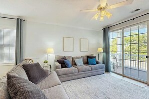 Ein Wohnzimmer mit einer grauen Eckcouch, zwei Beistelltischen mit Lampen, einem Deckenventilator und einer großen Schiebeglastür, die auf einen Balkon führt.