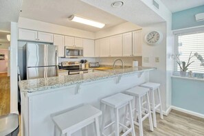 Eine moderne Küche mit weißen Schränken, einem Edelstahlkühlschrank, einer Mikrowelle, einem Toaster und einem Spülbecken. An der Kücheninsel stehen drei weiße Barhocker.