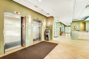 Eine Hotelhalle mit einer Rezeption, zwei Aufzügen, einem Selbstbedienungskiosk und einem braunen Teppich.
