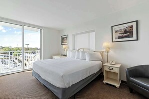 Ein Hotelzimmer mit einem großen Bett, zwei Nachttischen, einem Sessel, einem Balkon mit Blick und zwei gerahmten Bildern an der Wand.