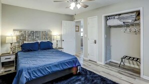 Un dormitorio con cama, lámparas de noche, ventilador de techo, vestidor y una puerta que da a otra habitación.