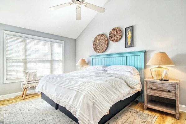 A bedroom with a bed, a ceiling fan, a window with blinds, a bedside table with a lamp, and a wall with decorative items. - Twelve Oaks Unit 108-4 (Pawleys Island)