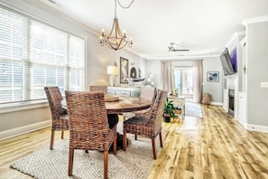 A dining area with a round table and wicker chairs, a chandelier, and a television on a stand. - Twelve Oaks Unit 108-4 (Pawleys Island)