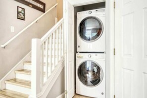 A laundry room with two stacked front-loading washing machines, a white staircase with wooden steps, and a white door. - Twelve Oaks Unit 108-4 (Pawleys Island)