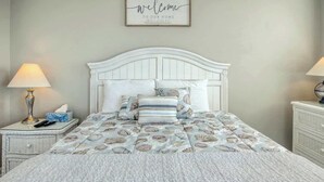 A bedroom with a white bed, a 'welcome to our home' sign, and a lamp on each side. - PacMan Get Away Condo (North Myrtle Beach)