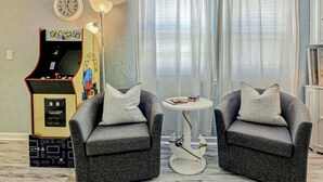A living room with two grey armchairs, a small round table, a Pac-Man arcade machine, a clock, and a lamp. - PacMan Get Away Condo (North Myrtle Beach)