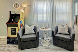 Ein Wohnzimmer mit zwei grauen Sesseln, einem kleinen runden Tisch, einer Pac-Man-Spielmaschine, einer Uhr und einer Lampe.