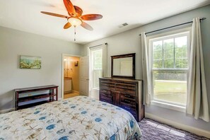 Ein Schlafzimmer mit einem Bett, einem Schminktisch, einem Deckenventilator, einem Fenster mit Vorhängen und einer Tür zu einem anderen Raum.