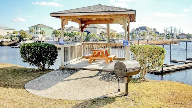 A gazebo with a picnic table and grill, overlooking a body of water with a dock and houses in the background. - Plantation Harbour G-16 (Little River)
