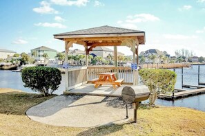 A gazebo with a picnic table and grill, overlooking a body of water with a dock and houses in the background. - Plantation Harbour G-16 (Little River)