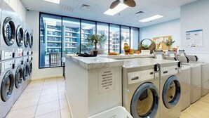 A laundry room with multiple washing machines, a control panel, and a sign indicating laundry room hours. - Renaissance Tower 211 (Myrtle Beach)