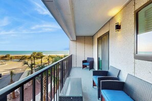 Uma varanda com vista para a praia, uma área de estar com cadeiras e uma mesa, e um luminário fixado na parede.