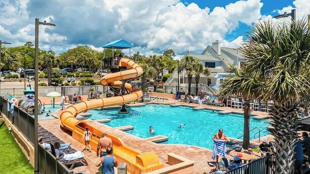 A swimming pool with a water slide, surrounded by palm trees and lounge chairs. - Caribbean Resort 525 (Myrtle Beach)