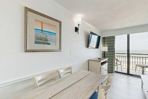 A room with a dining table, chairs, a television mounted on the wall, a small cabinet, and a balcony with a view of the sea. - Caribbean Resort 525 (Myrtle Beach)