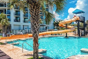 A swimming pool with a water slide, palm trees, and a multi-story building in the background. - Caribbean Resort 525 (Myrtle Beach)